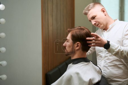 Photo for Diligent barber styling hair of his client who wearing cutting hair cape in modern barbershop - Royalty Free Image