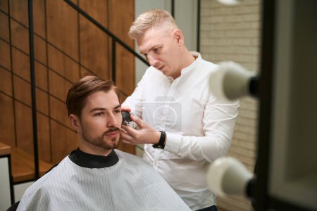 Foto de Peluquero caucásico cortando el pelo de su cliente que llevaba capa de corte de pelo en el interior - Imagen libre de derechos