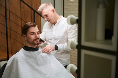 Photo for Photo of handsome hairdresser cutting hair of smiling man by using barber machine indoors - Royalty Free Image