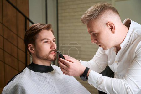 Téléchargez les photos : Coiffeur caucasien taille barbe de beau visiteur en utilisant un rasoir dans la salle de salon moderne - en image libre de droit