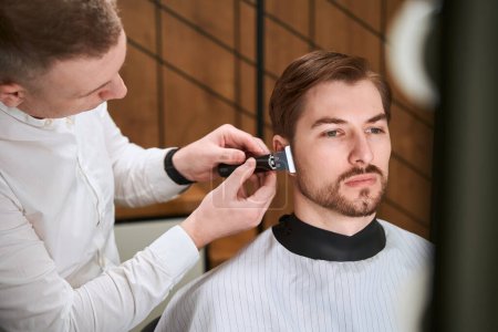 Photo for Concentrated hairdresser providing services to his client by using razor in modern room - Royalty Free Image