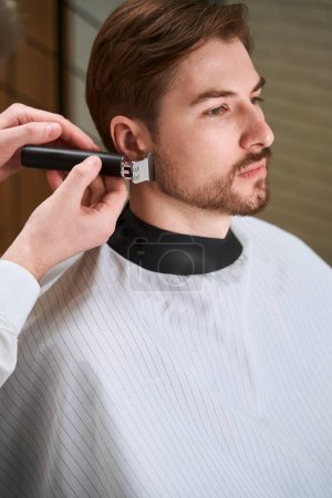 Photo for Hands of barber shaving beard of handsome man who wearing in hairdressing peignoir by using razor indoors - Royalty Free Image