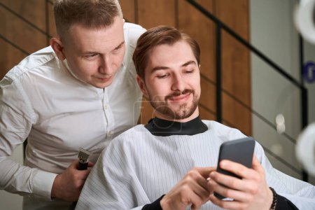 Photo for Male hairdresser and smiling man in cutting hair cape browsing cell phone in modern salon - Royalty Free Image