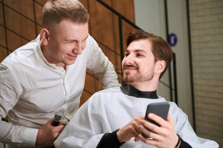Photo for Male visitor showing mobile and looking at barber while he browsing mobile indoors - Royalty Free Image