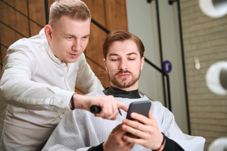 Photo for Enthusiastic barber and handsome man in hairdressing peignoir browsing cell phone in modern room - Royalty Free Image