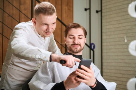 Photo for Professional hairdresser and young man in hairdressing peignoir browsing cell phone in modern barbershop - Royalty Free Image