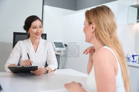 Photo for Brunette esthetician consults a blonde woman in a cosmetology clinic, she selects options for non-surgical skin tightening - Royalty Free Image