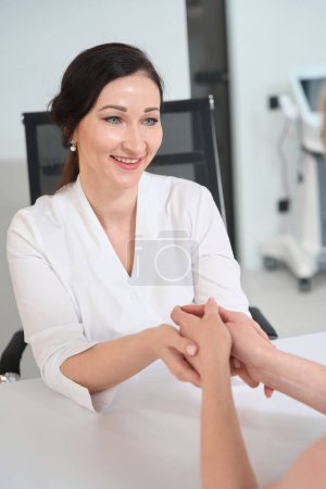 Foto de Sonriente morena cogida de la mano de una mujer sentada enfrente, esteticista en uniforme - Imagen libre de derechos