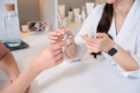 Foto de El doctor cosmetólogo sostiene una jeringa con una sustancia rejuvenecedora en sus manos, un cliente se sienta enfrente - Imagen libre de derechos