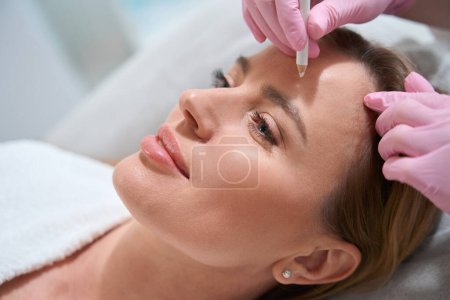 Foto de Esteticista utiliza un marcador especial para marcar t áreas de trabajo en la cara del paciente, la mujer se encuentra en el sofá cosmético - Imagen libre de derechos