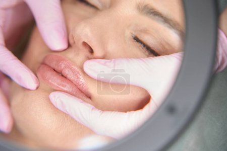 Foto de Especialista cosmetólogo examina el contorno de los labios de los pacientes a través de una lupa, una mujer después de inyecciones de belleza - Imagen libre de derechos