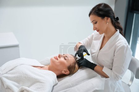 Foto de Morena esteticista realiza un procedimiento de inyección en una clínica de cosmetología, el paciente se acuesta en el sofá - Imagen libre de derechos