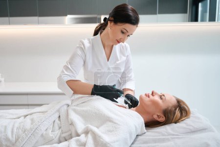 Téléchargez les photos : Esthéticienne dans une clinique de cosmétologie fait une injection dans le cou d'une femme, la patiente s'allonge sur le canapé - en image libre de droit