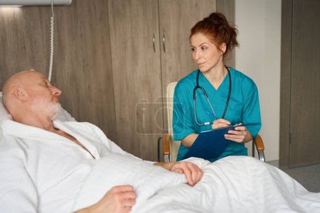 Foto de Doctora de uniforme sentada con portapapeles cerca del paciente que se acuesta en la cama y cuenta su diagnóstico en la sala de hospital - Imagen libre de derechos