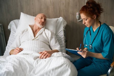 Téléchargez les photos : Médecin féminin avec presse-papiers écrivant des symptômes et prescrivant un nouveau médicament au patient masculin au lit dans une chambre d'hôpital - en image libre de droit