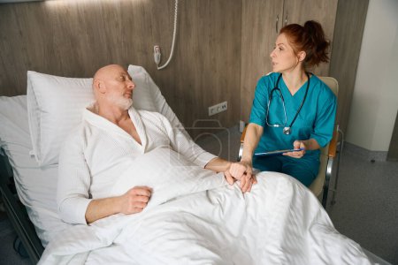 Téléchargez les photos : Médecin féminin en uniforme assis près du patient masculin au lit et le soutenant tout en parlant de récupération rapide en chambre à l'hôpital - en image libre de droit