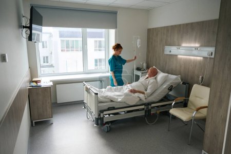 Foto de Médico femenino visitando paciente varón enfermo con gotero que yace en la cama del hospital después de la cirugía - Imagen libre de derechos