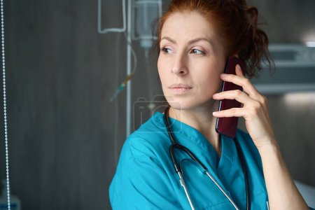 Photo for Concentrated female doctor with stethoscope talking on the smartphone in chamber at clinic - Royalty Free Image