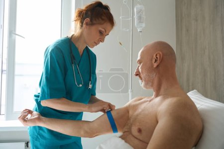 Téléchargez les photos : Assistant de laboratoire serrant le garrot médical sur le bras du patient avant de prélever un échantillon de sang dans la chambre de l'hôpital - en image libre de droit