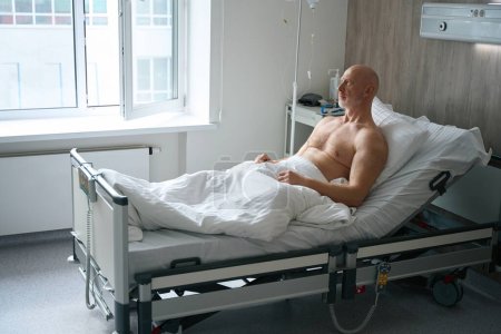 Téléchargez les photos : Malade homme mûr se reposant au lit et se rétablissant après une chirurgie médicale dans la salle d'hôpital - en image libre de droit