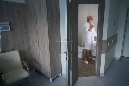 Foto de Enfermo hombre maduro en bata blanca de pie cerca del fregadero y hacer asuntos de rutina en el baño de la cámara del hospital - Imagen libre de derechos