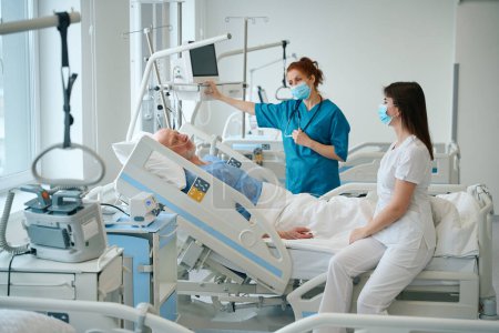Téléchargez les photos : Médecins féminins masqués rendant visite à leur patient masculin allongé dans le lit de la salle d'hôpital - en image libre de droit