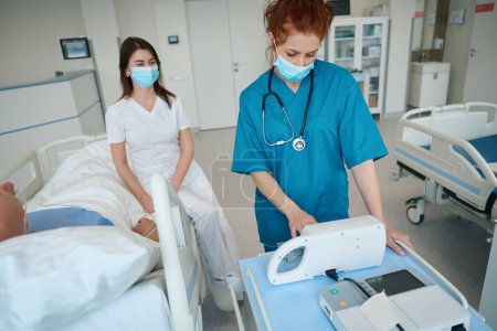 Téléchargez les photos : Infirmière en masque médical personnalisant la pompe à seringue pour administrer des médicaments pendant que le médecin communique avec le patient allongé sur le lit dans l'unité de soins intensifs - en image libre de droit