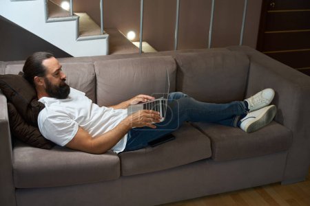Foto de Hombre de mediana edad se encuentra en un sofá con un ordenador portátil, que está situado cómodamente en la zona de recreo - Imagen libre de derechos