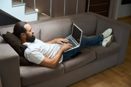 Foto de Hombre de mediana edad está descansando en un sofá con un ordenador portátil, que está situado cómodamente en la zona de recreo - Imagen libre de derechos