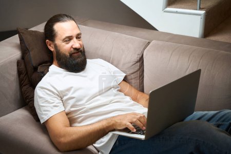 Téléchargez les photos : L'homme satisfait se repose sur un canapé avec un ordinateur portable, il est confortablement situé dans la zone de loisirs - en image libre de droit