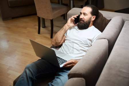 Téléchargez les photos : Homme avec une barbe bien entretenue utilise ordinateur portable et téléphone mobile pour communiquer, il est confortablement situé dans la zone de loisirs - en image libre de droit