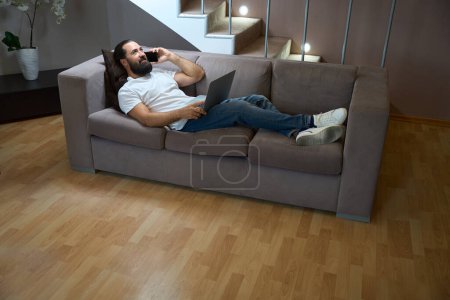 Photo for Hotel guest uses a mobile phone and a laptop to communicate, a man comfortably sits on a soft sofa - Royalty Free Image