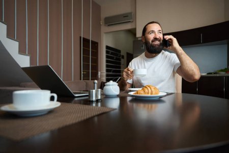 Foto de Hombre alegre bebe café y se comunica emocionalmente por teléfono, él está sentado en una mesa en el área de la cocina - Imagen libre de derechos
