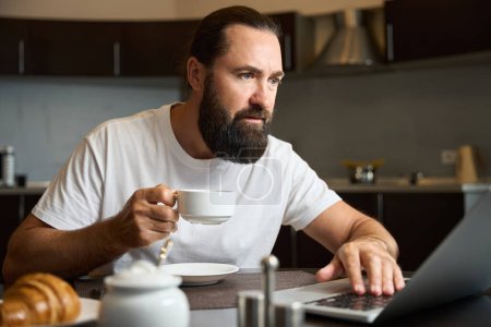 Photo for Worried male is drinking coffee with a croissant and working on a laptop, he is sitting in the kitchen area - Royalty Free Image