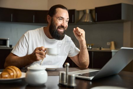 Photo for Bearded man drinks coffee and communicates emotionally online, he is seated at a table in the kitchen area - Royalty Free Image
