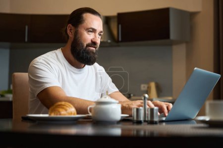 Foto de El hombre de negocios se comunica por computadora portátil durante el desayuno, la mesa está puesta para una persona - Imagen libre de derechos