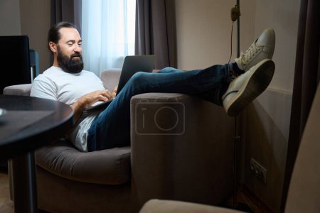 Photo for Middle-aged male is located on the sofa with a laptop, he is in comfortable casual clothes - Royalty Free Image