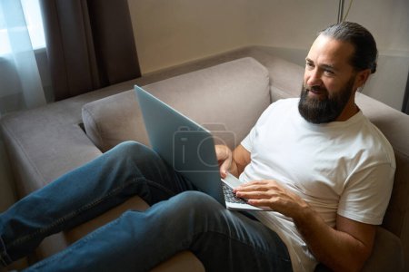 Foto de Varón barbudo en jeans se sienta en una silla cómoda con un ordenador portátil, se comunica en línea - Imagen libre de derechos