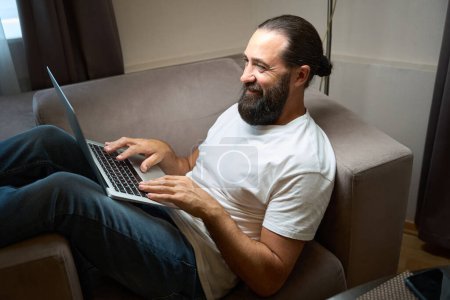 Foto de Hombre barbudo en jeans se sienta en una silla cómoda con un ordenador portátil, se comunica en línea - Imagen libre de derechos