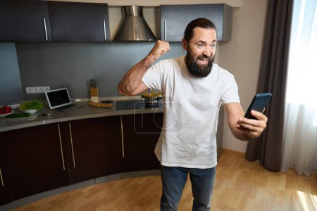 Foto de Huésped del hotel se comunica emocionalmente en línea en un teléfono móvil, él está en el área de cocina de la habitación de hotel - Imagen libre de derechos