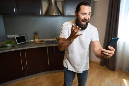 Foto de Hombre adulto se comunica emocionalmente en línea en un teléfono móvil, él está en el área de la cocina de la habitación de hotel - Imagen libre de derechos