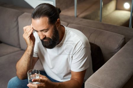 Photo for Bearded male holds a glass of water in his hands, he suffers from a headache - Royalty Free Image