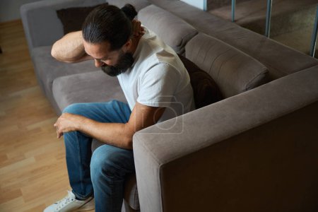 Photo for Bearded man is holding on to his sore neck, he is sitting on a soft sofa - Royalty Free Image