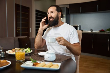 Foto de El barbudo está hablando por teléfono en el desayuno, los platos con comida están sobre la mesa. - Imagen libre de derechos