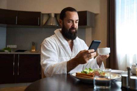 Photo for Male in a bathrobe sits at a table with a phone and breakfast, a plate of food is on table - Royalty Free Image