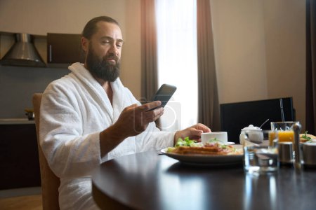 Foto de El hombre en un albornoz se sienta en una mesa con un teléfono y el desayuno, un plato de comida está en la mesa - Imagen libre de derechos
