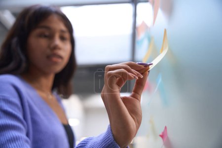 Téléchargez les photos : Femme d'affaires avec visage flou collant avis papier sur le mur de verre tout en se tenant à l'intérieur - en image libre de droit