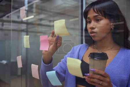 Téléchargez les photos : Femme d'affaires en vêtements fantaisie tenant tasse de café et collant des notes colorées tout en se tenant devant le mur de verre à l'intérieur - en image libre de droit