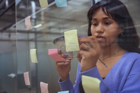 Foto de Mujer atractiva con taza de café en la mano escrito en papel de aviso en la pared de cristal en la oficina moderna - Imagen libre de derechos