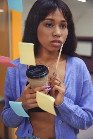 Photo for Pretty female in fancy clothes lost in thought while holding cup of coffee and pencil indoors - Royalty Free Image